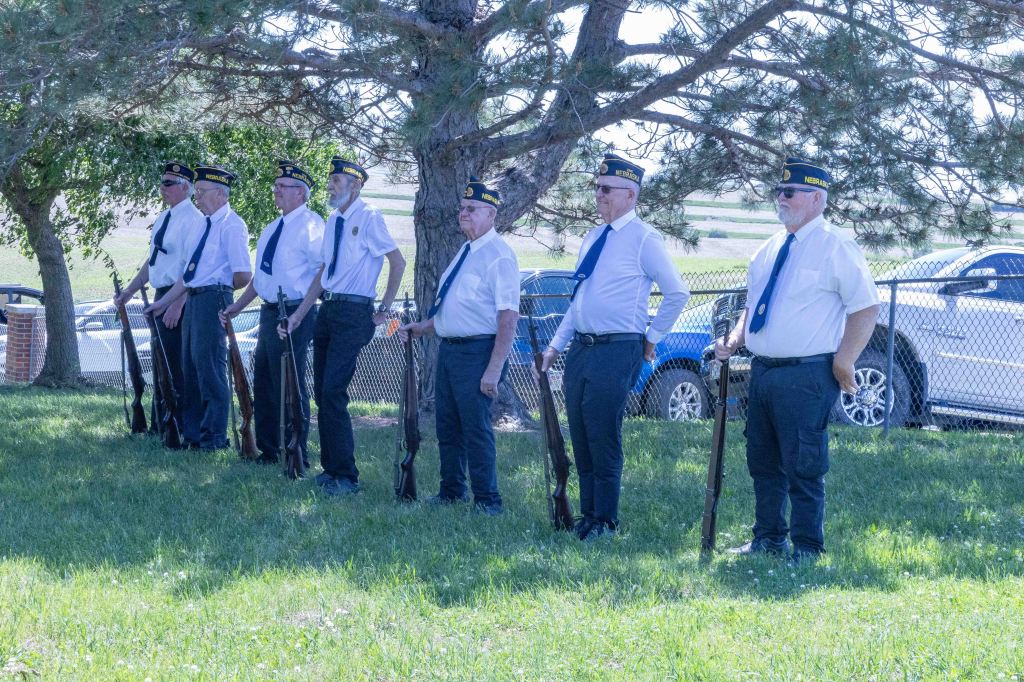 A group of veterans in uniform standing in a line, holding rifles, beneath a tree during a ceremony.