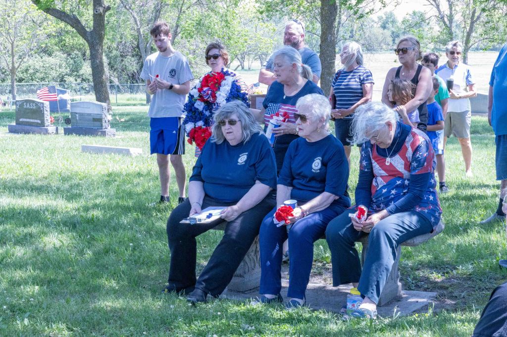 A group of people, including several women in blue shirts and one in a patriotic outfit, gathered at a cemetery for a memorial event. Some attendees hold flowers or drinks while sitting on a stone bench amidst a grassy area with gravestones in the background.