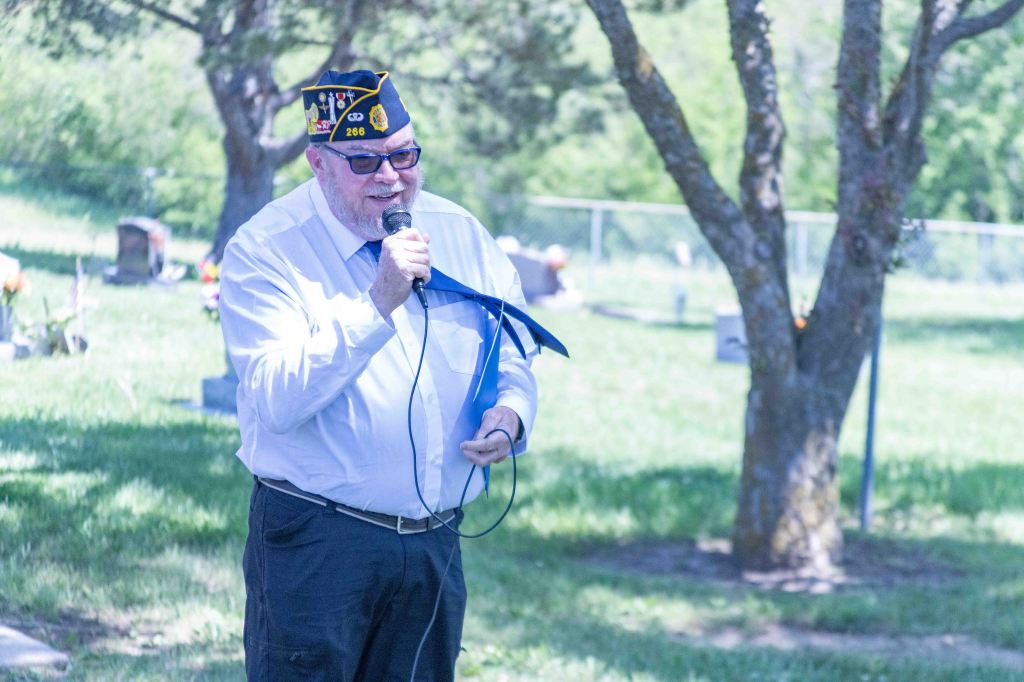 A man in a veterans' cap speaking into a microphone at a cemetery, surrounded by trees and graves.