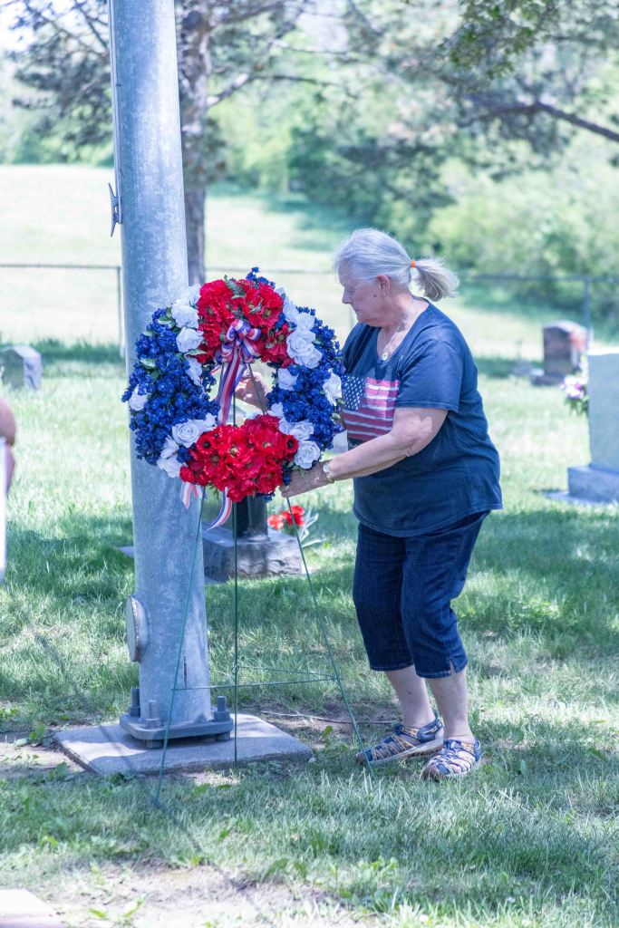 An elderly woman arranging a red, white, and blue floral wreath at a cemetery, with green grass and gravestones in the background.