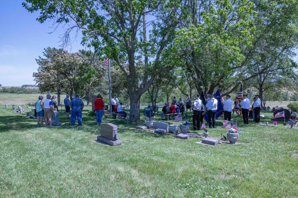 A group of people gathered in a cemetery for a memorial service, with American flags placed on gravestones and trees in the background.