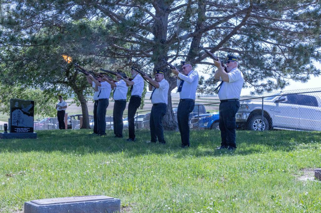 A group of six uniformed individuals participates in a ceremonial gun salute, firing rifles towards the sky in front of a gravestone, with greenery and a tree in the background.