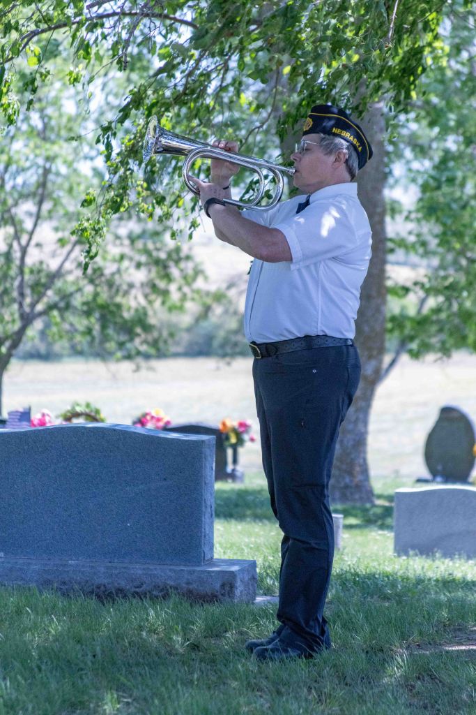 A man in military attire plays the trumpet at a cemetery, surrounded by gravestones and greenery.