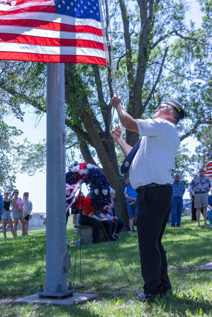 A man in military attire raises the American flag at a memorial event, surrounded by attendees in a park-like setting.
