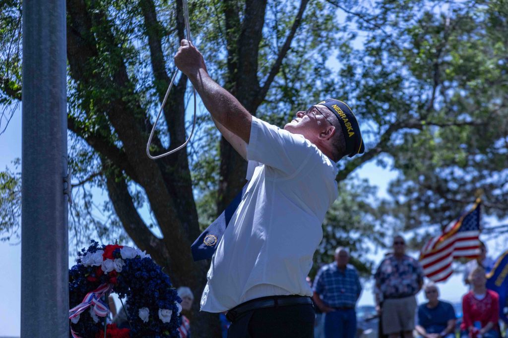 A man in a white shirt and a Nebraska cap raises a flag at a memorial ceremony, with a wreath in the foreground and an audience in the background.