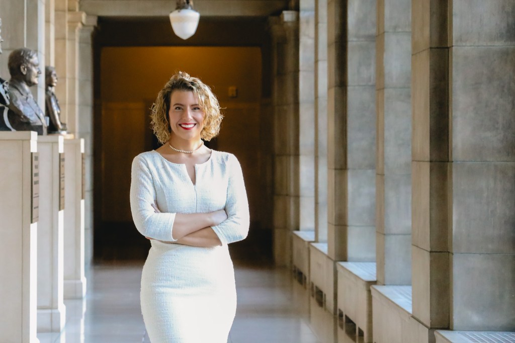 A confident woman with curly hair stands in a corridor, wearing a white dress and smiling, with classical sculptures and stone columns in the background.
