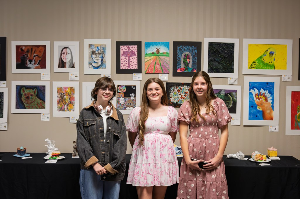Three young women stand in front of a colorful art display featuring various student artworks on the wall. They are smiling and wearing different styles of dresses. Table with art pieces in the foreground.