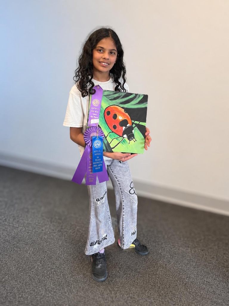 A young girl with curly hair is smiling while holding a colorful painting of a ladybug. She is wearing a white t-shirt and flared jeans, and she has a purple ribbon indicating a first place award for fine arts.