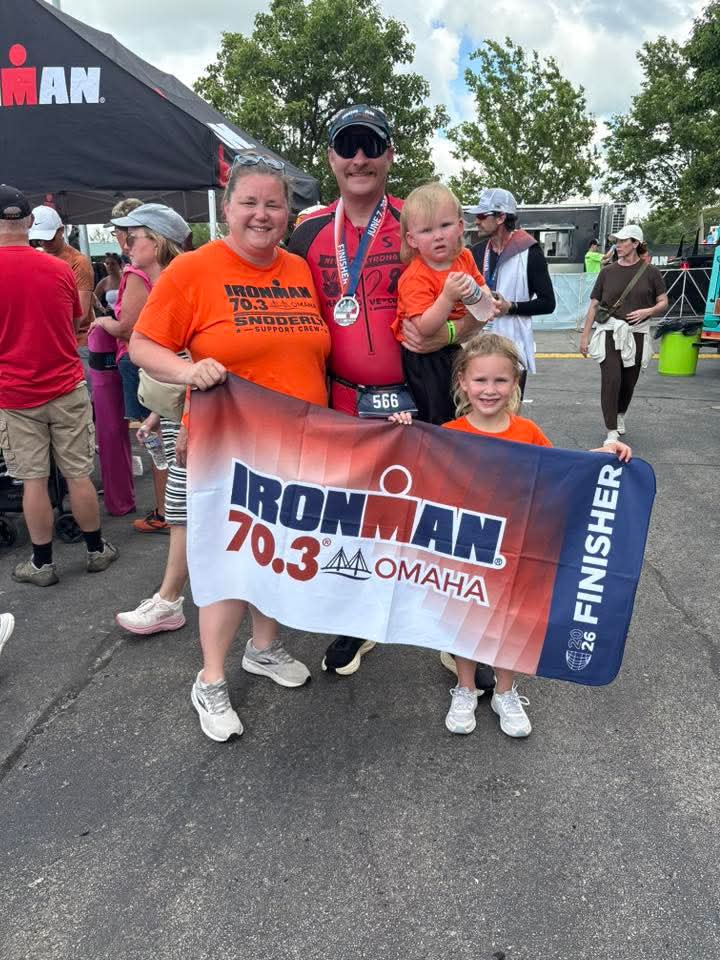 A family poses together after an Ironman 70.3 event in Omaha, holding a finisher banner. The group includes a man wearing a medal, a woman, and two children, all in matching orange shirts.