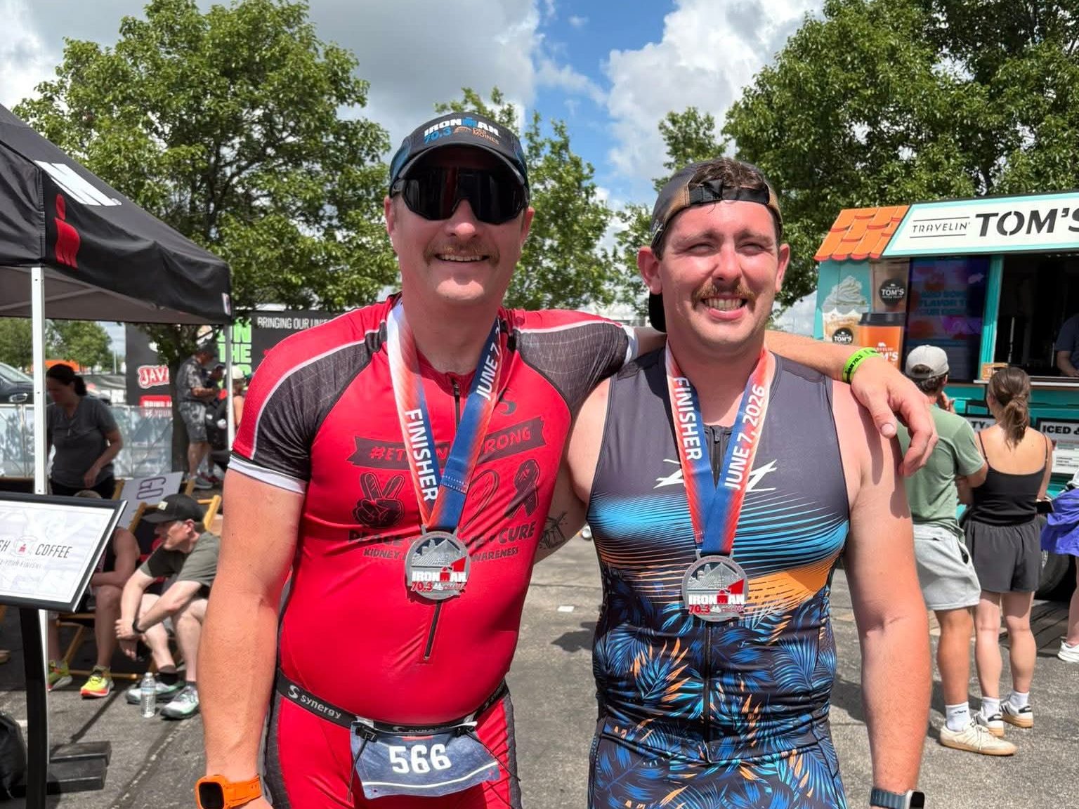 Two male athletes smiling and posing together after completing a race, wearing finisher medals and athletic gear, with a food truck and spectators in the background.