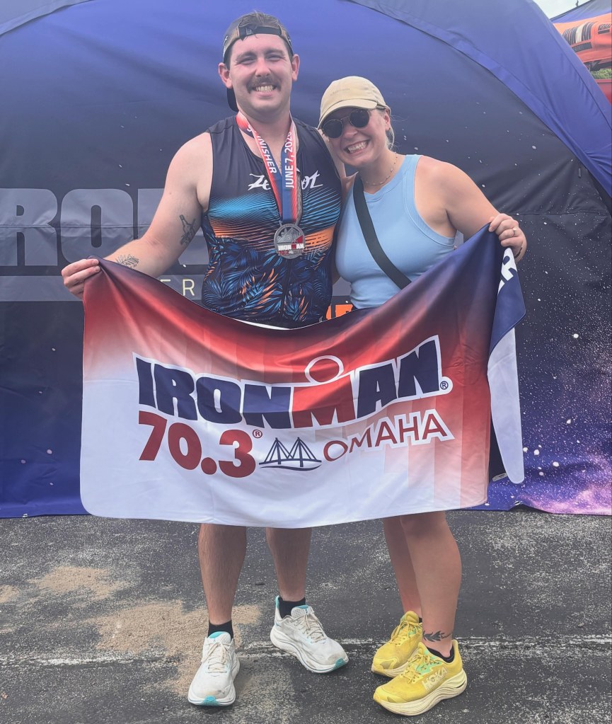 A man wearing a tank top and a medal is standing next to a woman holding an 'Ironman 70.3 Omaha' banner, both smiling in front of a dark blue backdrop.