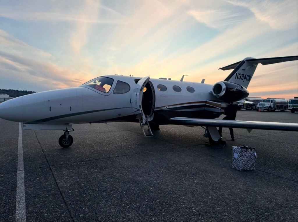 A private jet is parked on an airport tarmac at sunset, with its door open and a suitcase nearby.