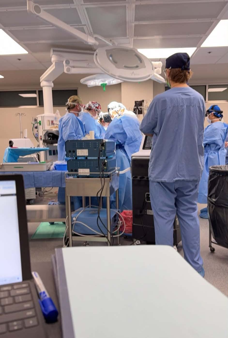 A surgical team in blue scrubs performing a procedure in an operating room, with surgical equipment and monitoring devices visible. The scene also shows a computer setup in the foreground.