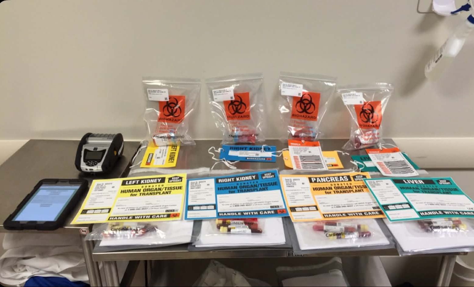 A medical table displaying various organ transplant bags labeled with biohazard symbols, including left kidney, right kidney, pancreas, and liver, along with a tablet and labeling equipment.