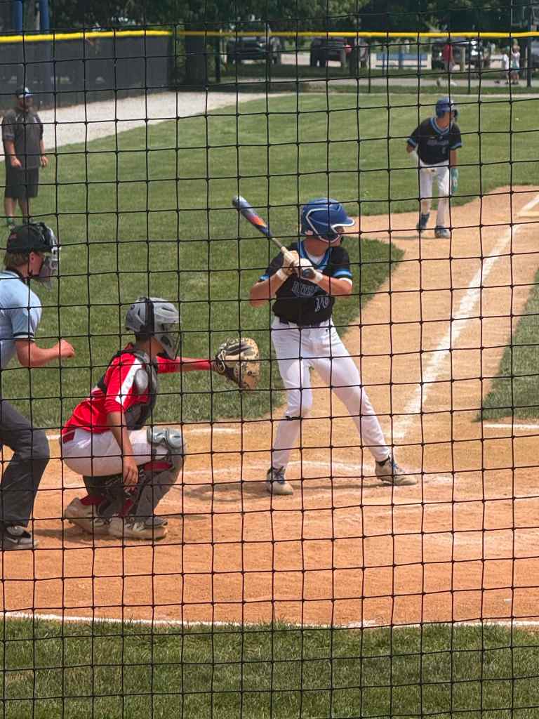 A young baseball player in a blue jersey stands ready to bat at home plate, with a catcher in red gear crouched behind him and an umpire nearby. Another player stands on the pitcher's mound in the background.