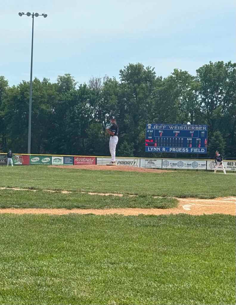 A baseball pitcher on the mound preparing to throw, with a scoreboard in the background showing game details at Lynn R. Pruess Field.