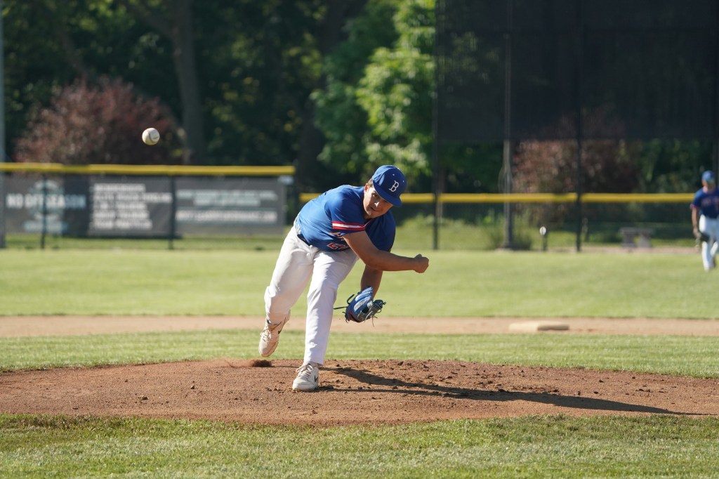 A baseball pitcher in a blue jersey throws a ball from the pitcher's mound during a game on a sunny day.