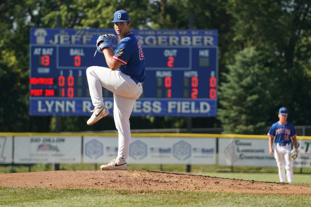 A pitcher in a blue baseball uniform winds up on the pitcher's mound, with a scoreboard visible in the background displaying game statistics.