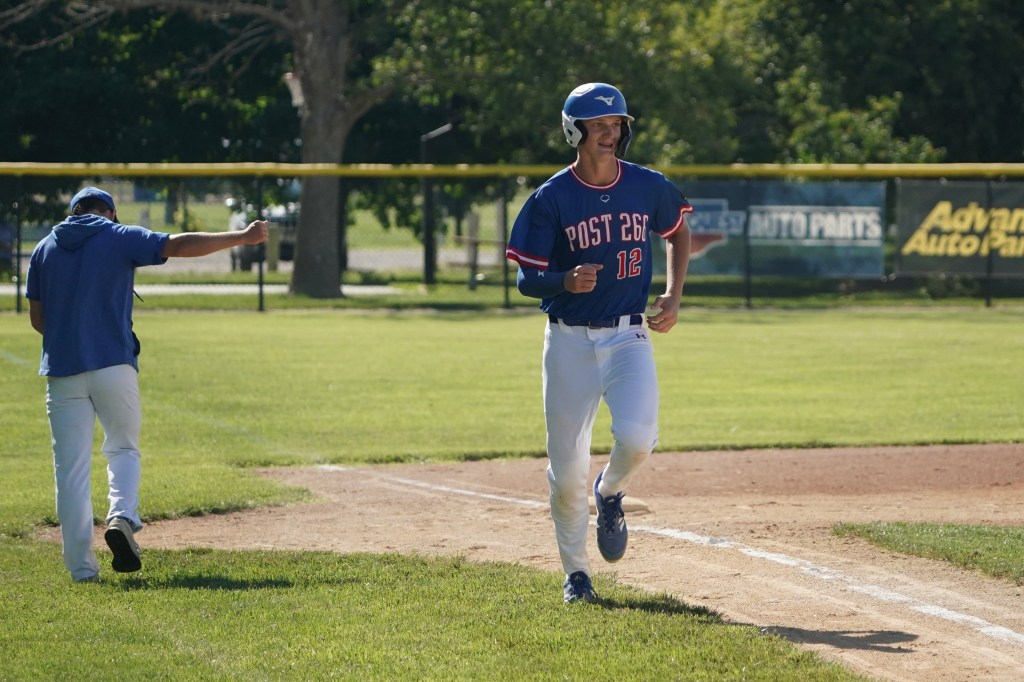 A baseball player in a blue jersey with 'Post 26' and number 12 smiles while running towards home plate on a sunny day. Another player in a blue hoodie stands in the background.