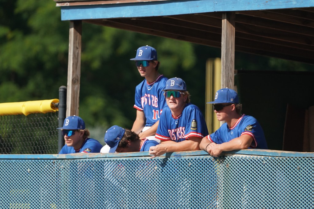 A group of young baseball players in blue uniforms with 'B' logos sitting in a dugout, watching the game.