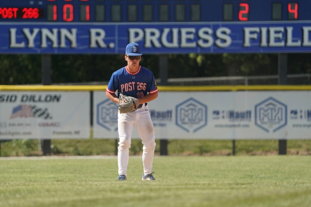 A young baseball player wearing a blue and red jersey with the number 16 stands in a field, looking down while holding a glove. In the background, the scoreboard shows a score of 2-4.