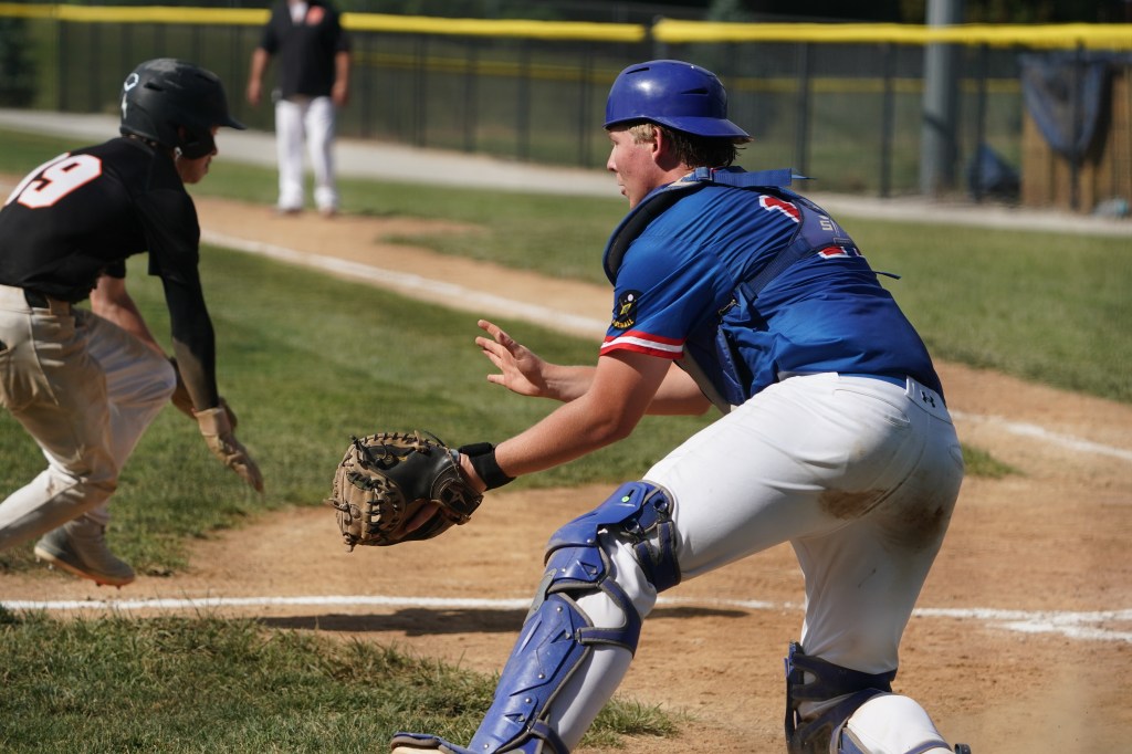 A youth baseball catcher in a blue uniform prepares to catch a pitch while a player in black runs towards the base.