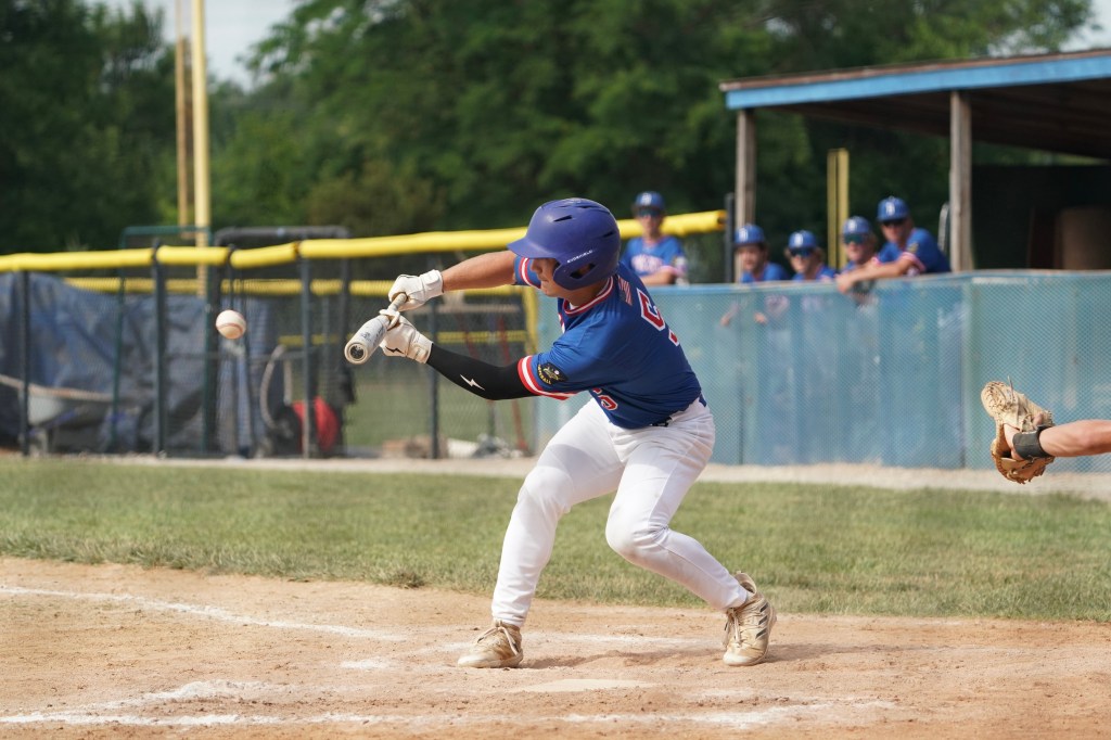 A young baseball player swings at a pitch, attempting to hit the ball during a game. He is wearing a blue jersey and white pants, with a batter's box visible in the foreground.
