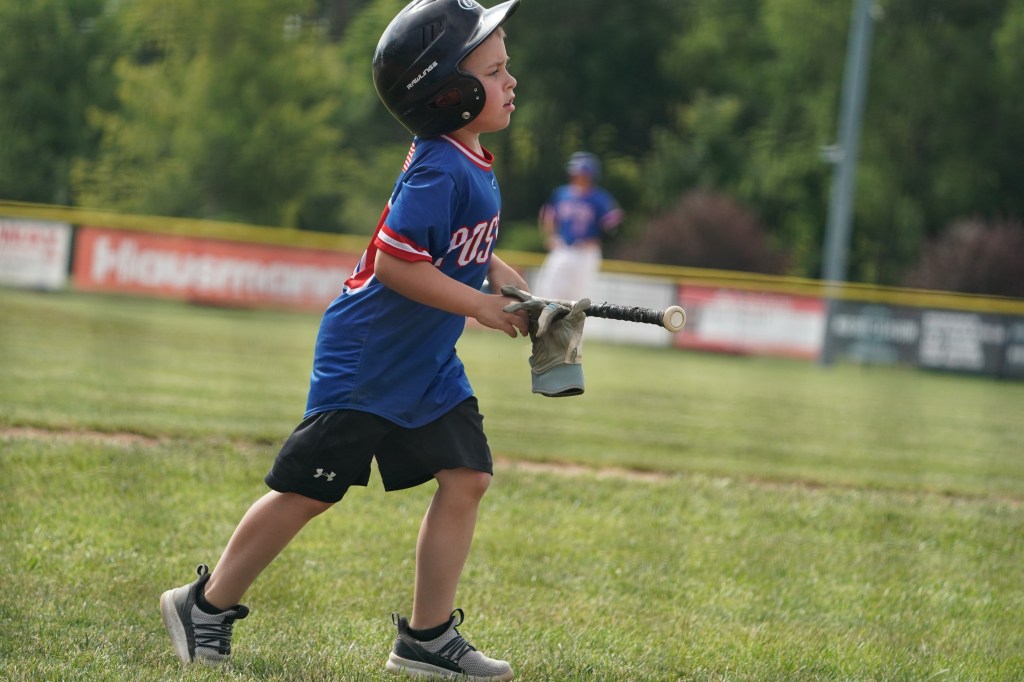A young boy wearing a baseball helmet and jersey runs on a grassy baseball field, holding a bat and a glove.