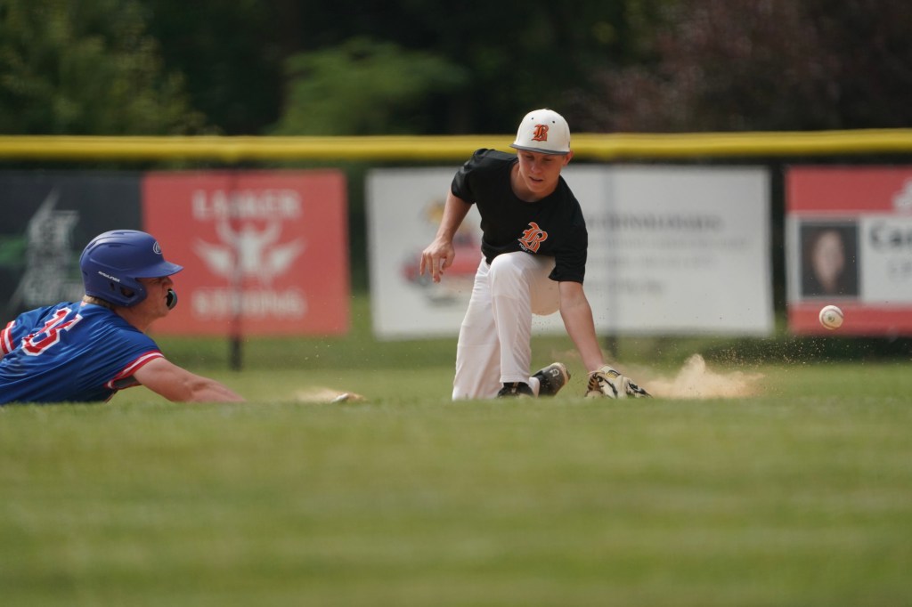 A baseball player in a black shirt is attempting to field a ground ball while another player in a blue shirt slides into base.
