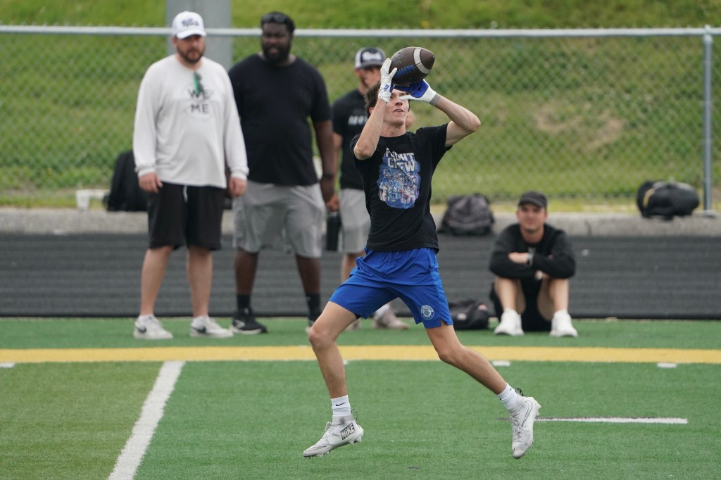 A young athlete catching a football during practice, with several spectators in the background.