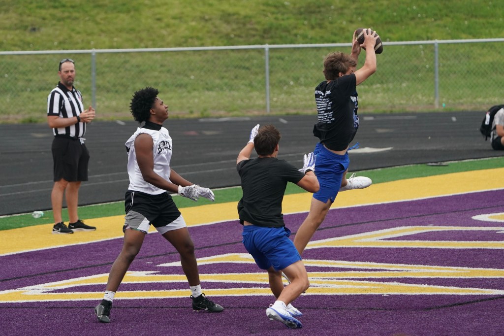 A football player jumps to catch a football while two other players defend, with a referee observing in the background.