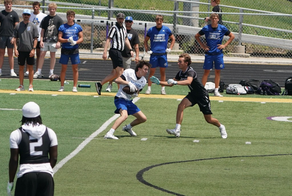 A high school football practice scene with two players in action. One player in a white shirt is running with the ball while being pursued by a player in black. In the background, several teammates and coaches are watching from the sidelines.