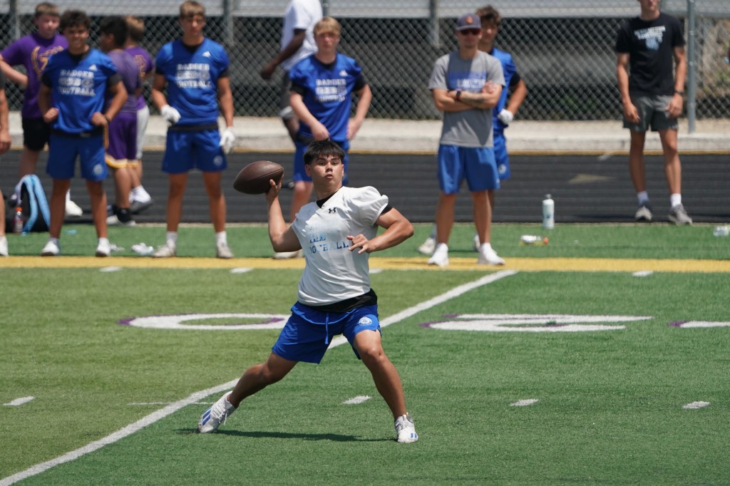 A young athlete throwing a football during a practice session on a green turf field, with several teammates observing in the background.
