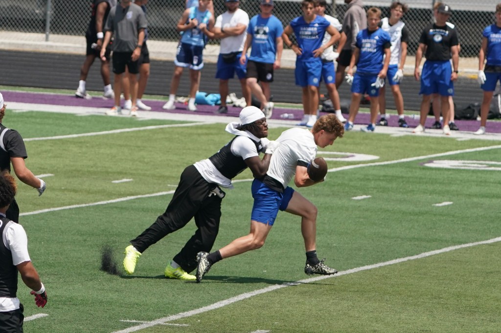 A football player in a white shirt and blue shorts is being tackled from behind by another player in a black shirt and pants during a practice session on a green field.