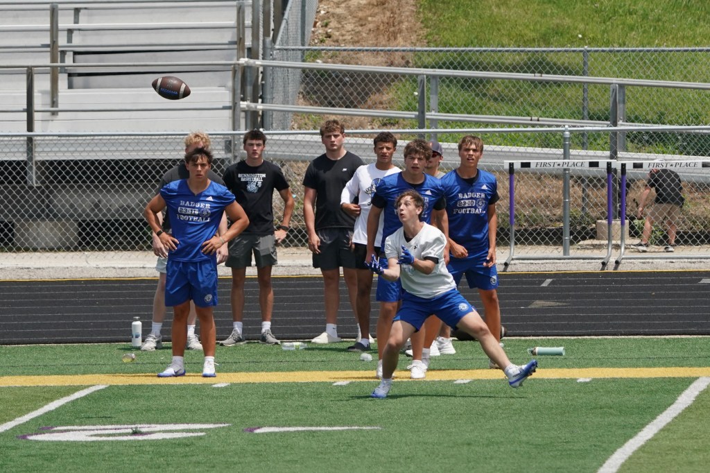 A group of young athletes watches as a teammate throws a football during a practice session on a sports field.