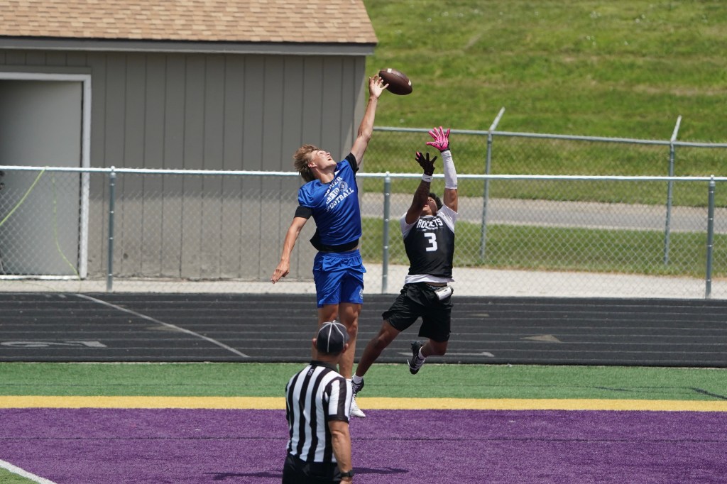 A football player in a blue jersey jumps high to catch a football while another player in a black jersey attempts to defend. A referee in the foreground observes the play.