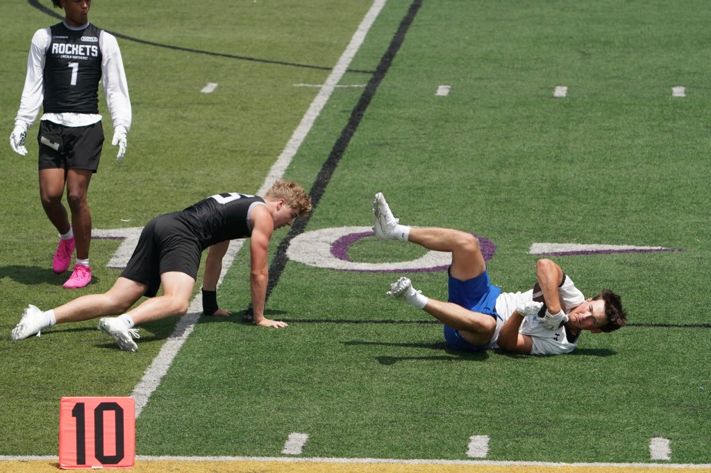 Two young male athletes on a football field; one player is down on the ground in pain while the other stands nearby.