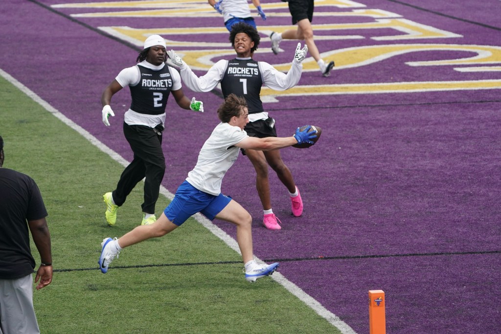 A player in a blue uniform catches a football while diving, with two teammates in black jerseys celebrating in the background on a purple football field.