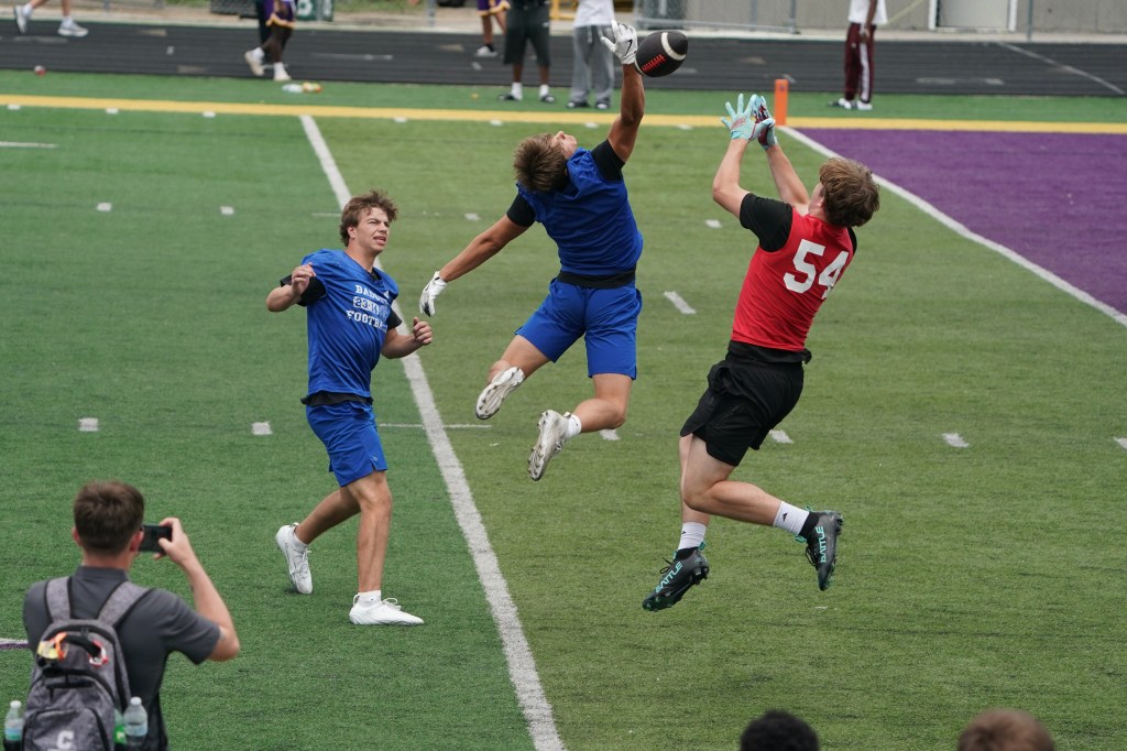 A football player in blue jumps to catch a football while another player in red attempts to block the pass during a practice on a grassy field. In the background, a small crowd observes the activity.