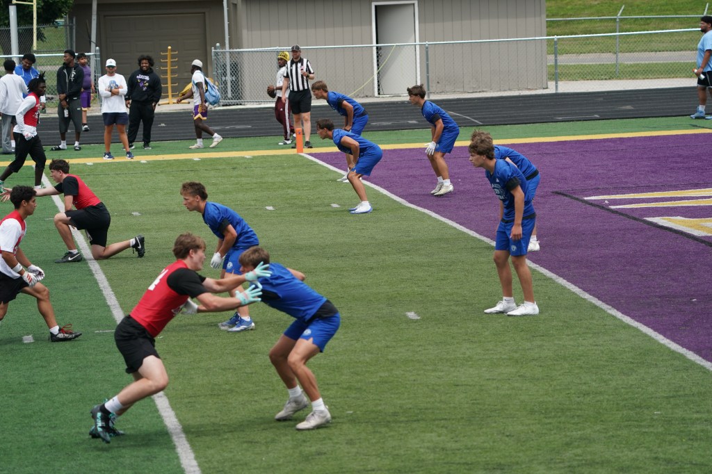 A group of young athletes in blue and red jerseys practicing on a football field. Some players are in a ready stance, while others observe from the sidelines.
