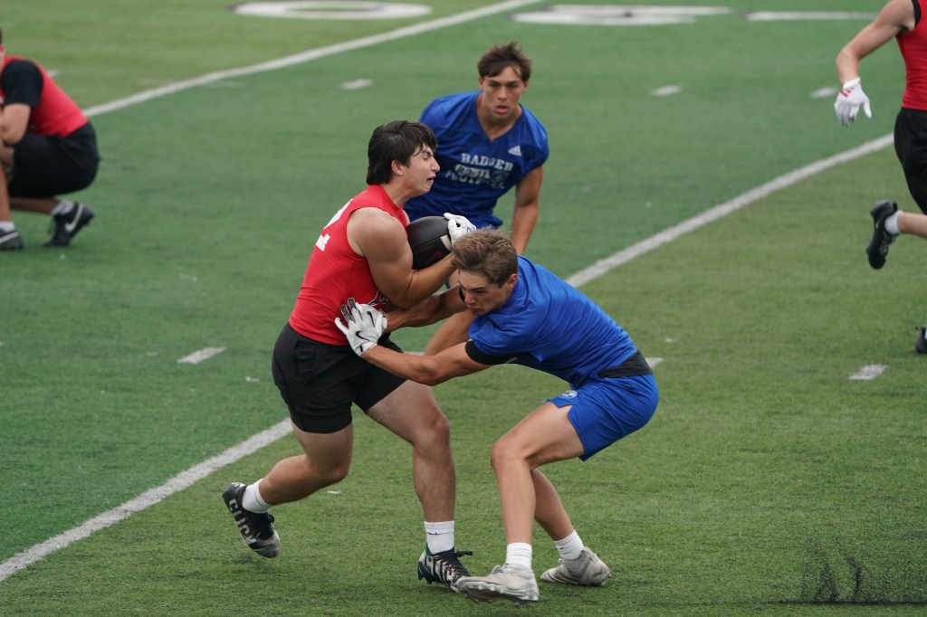 Two young male athletes engaged in a competitive tackle during a football practice on a green turf field. One player in a red jersey is holding the football, while the other in a blue jersey is attempting to make a tackle.