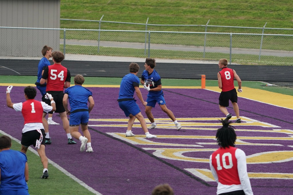 A group of young athletes participating in a football practice on a field with purple and yellow markings. Players are in various colored jerseys, with one player poised to throw the ball while others are in motion.