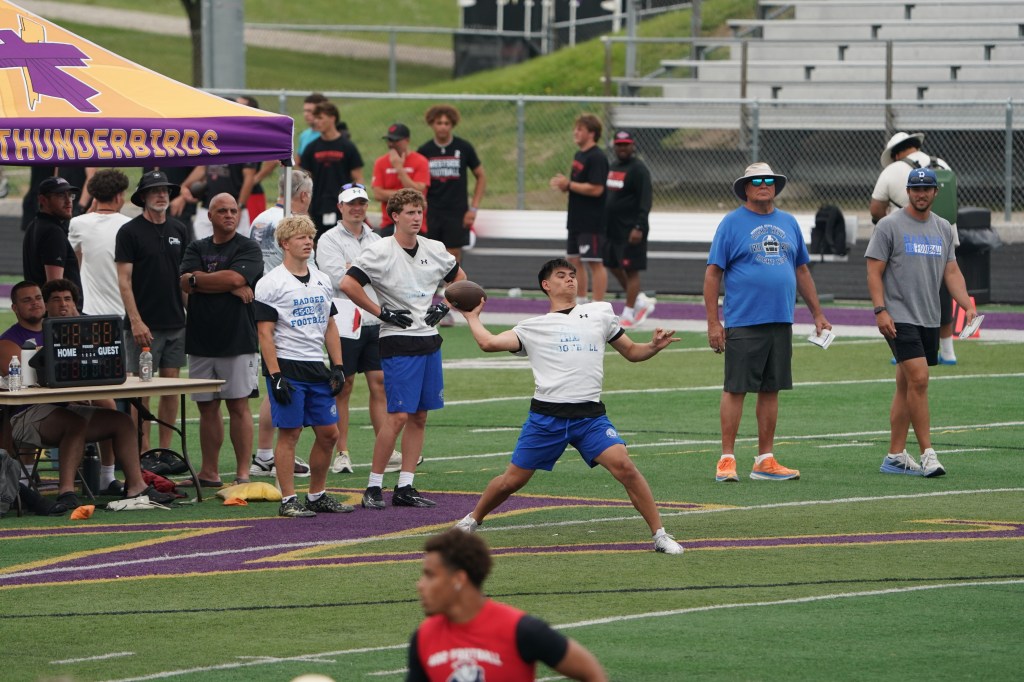A young athlete in a white shirt and blue shorts throws a football during practice, with coaches and other players observing from the sidelines under a purple tent labeled 'THUNDERBIRDS'.