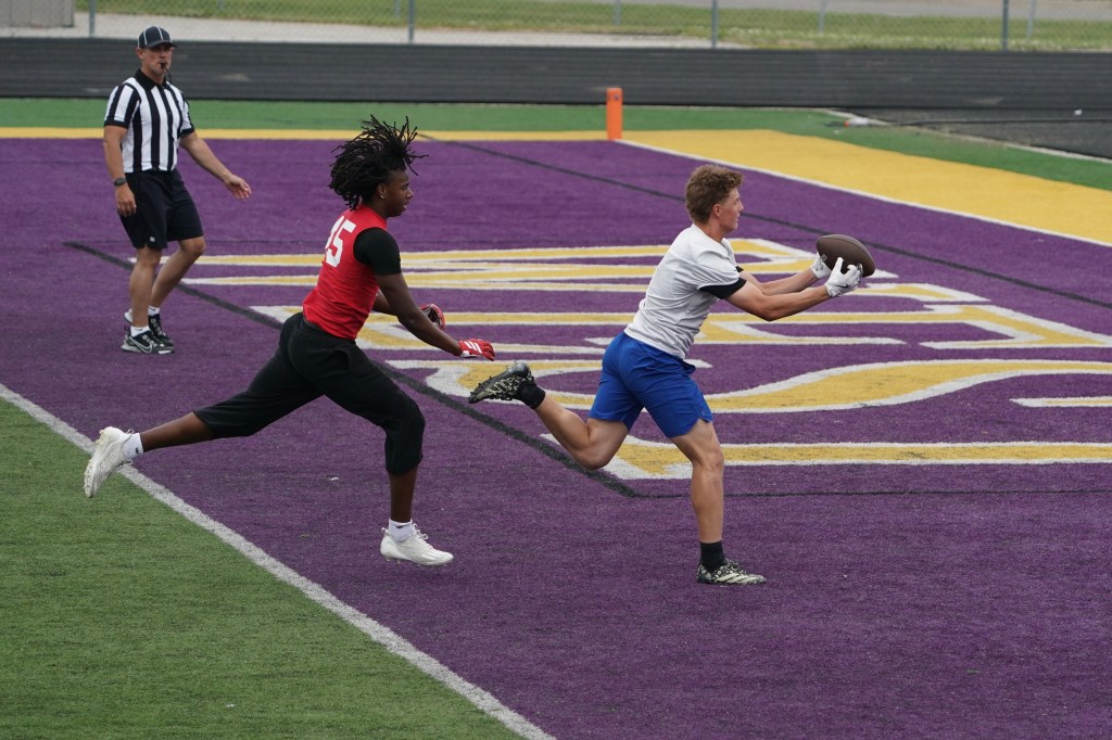 A football player in a white shirt catches a football while running on a purple field, with a defender in a red shirt sprinting behind him. A referee observes the play in the background.