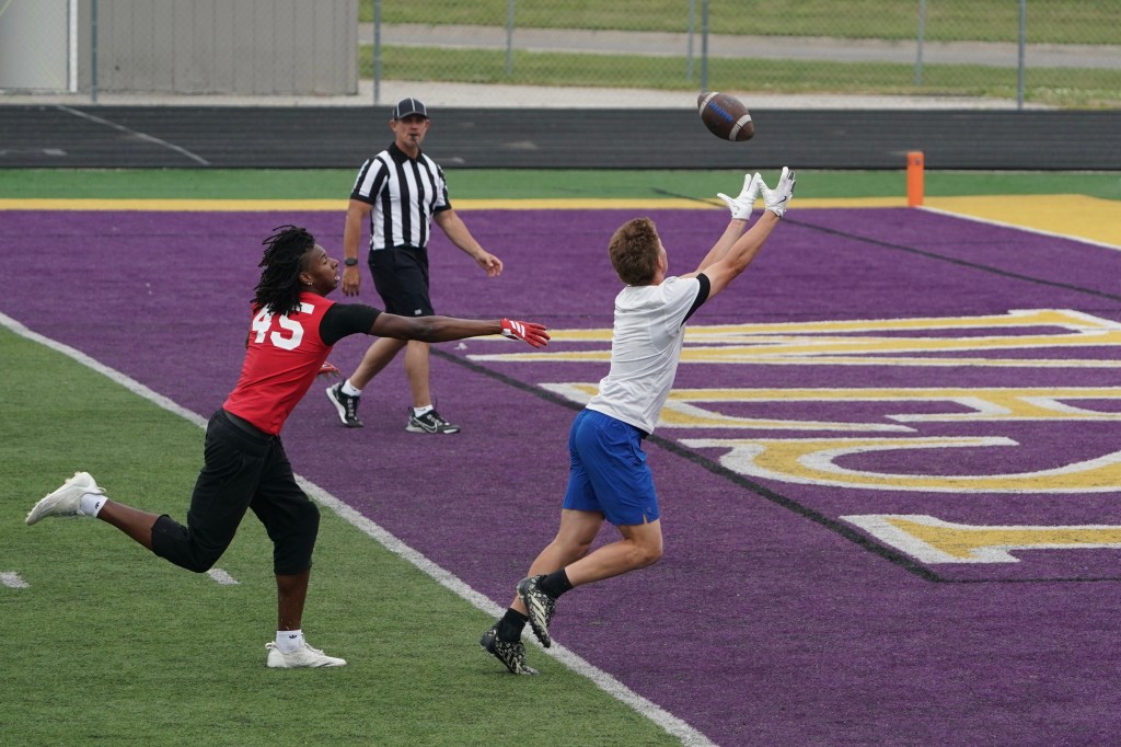 A football player in a white shirt catches a pass while a defender in a red shirt attempts to intercept. An official is observed in the background on the field with purple turf.