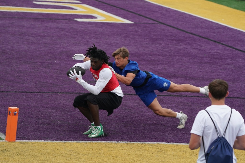 A football player in a red jersey catches the ball while being defended by another player in a blue jersey who is diving to tackle him. The scene takes place on a purple and yellow field.