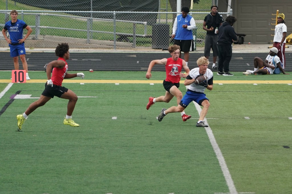 A group of young athletes playing football on a field, with one player wearing a white shirt running with the ball while being pursued by two others in red shirts.