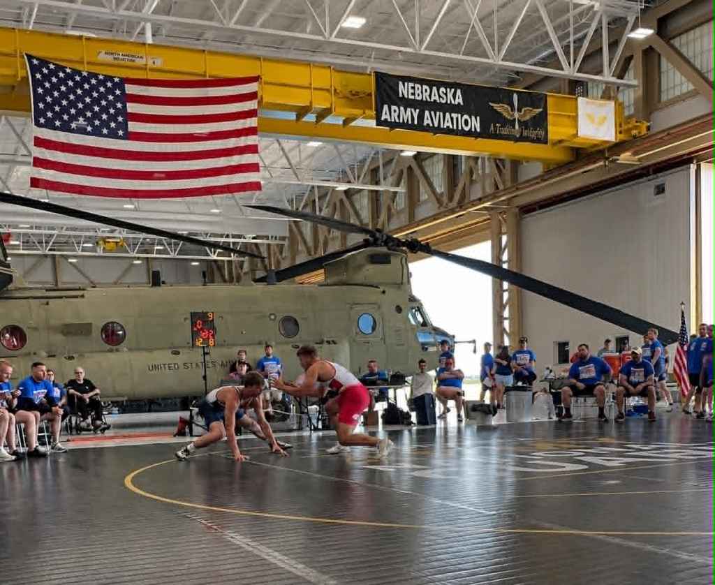 A wrestling match taking place inside a hangar, with a U.S. flag and a helicopter in the background. Two wrestlers compete on a mat while an audience watches.