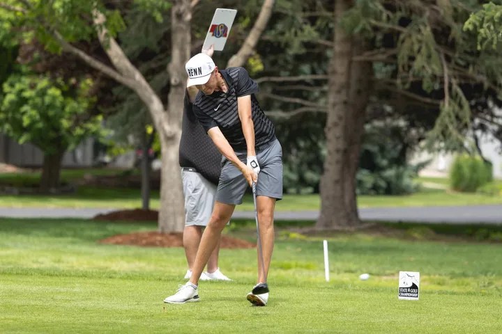 A golfer in a black and gray outfit swings a golf club on a sunny day, with a blurred figure holding a sign in the background and trees surrounding the scene.