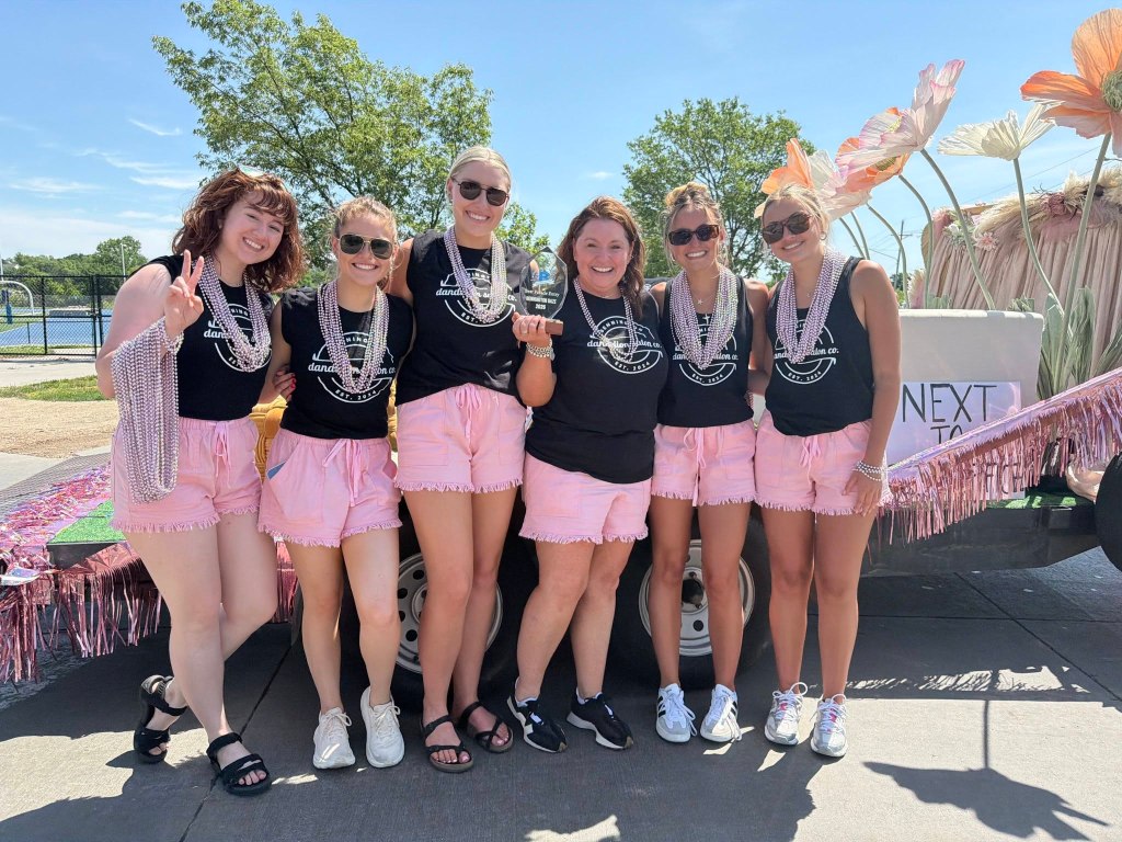 A group of six women standing together, wearing matching black shirts and pink shorts, adorned with pearl necklaces and smiling at the camera. They are posing in front of a decorated float with large floral decorations in a sunny outdoor setting.